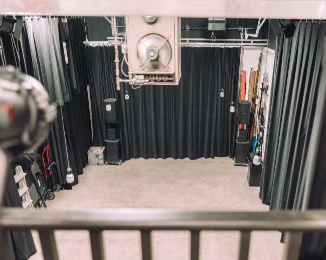 The image shows an empty stage area with black curtains, overhead lights, and various equipment arranged around the sides.