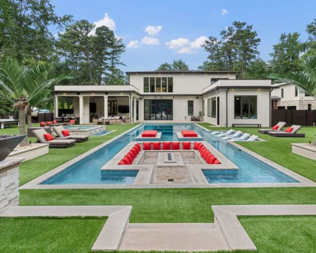 The image features a modern home with a symmetrical pool surrounded by lush greenery and lounge seating.