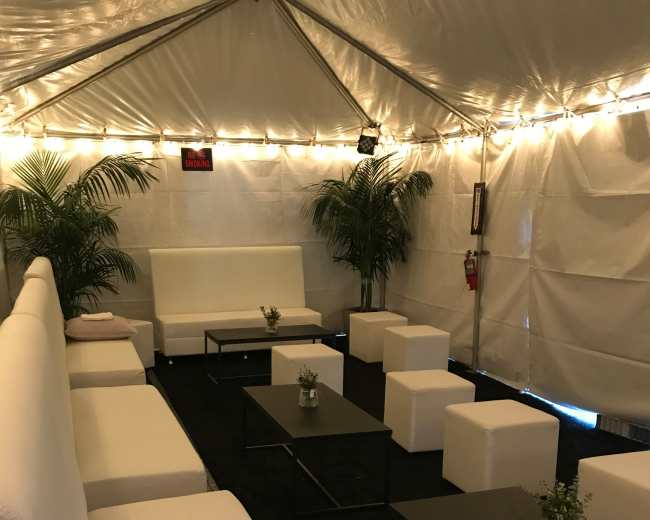 The image shows a white tent interior with several modern couches, low tables, and decorative plants arranged in a lounge setting.