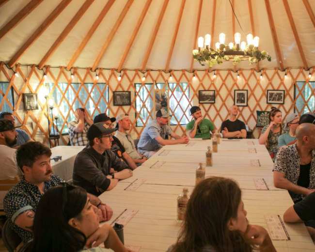 A group of people sits around a long table inside a yurt, with various photographs hanging on the walls.