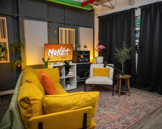 The image shows a cozy room with a yellow sofa, a light-colored chair, and a television displaying "Monset Talks," surrounded by plants and black curtains.
