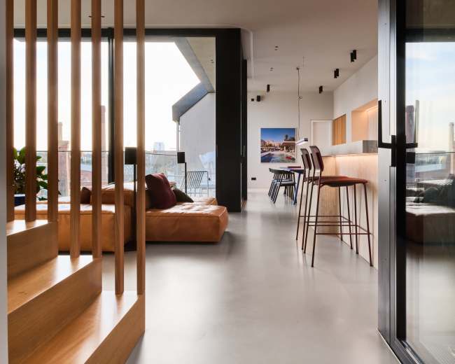 The image shows a modern interior space featuring a staircase, a leather sofa, and a bar with high stools, all illuminated by natural light from large windows.