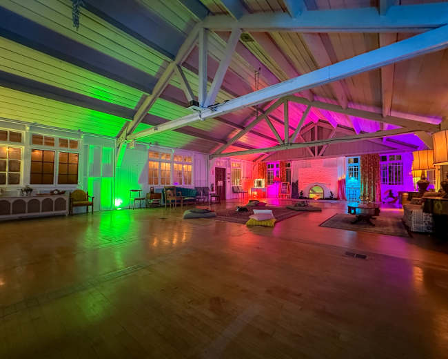 The image shows a spacious interior room with high wooden ceilings, featuring colorful lighting and various seating arrangements including cushions and chairs.