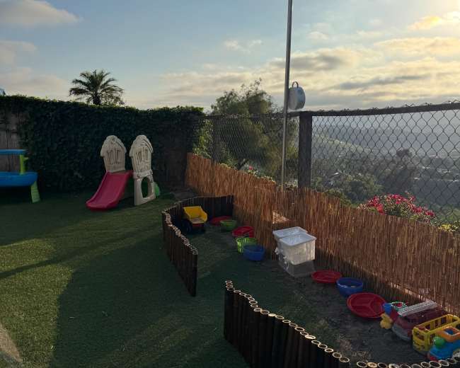 The image shows a children's play area featuring a slide, playhouses, and various toys, surrounded by a fence and decorated with green grass-like carpet and bamboo accents.