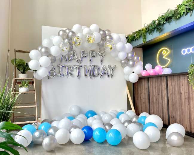 The image features a birthday decoration setup with a "HAPPY BIRTHDAY" banner beneath a balloon arch, surrounded by a mix of blue, silver, and white balloons on the floor.