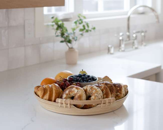 A basket filled with various baked goods and fresh fruit is placed on a white kitchen countertop near a window.
