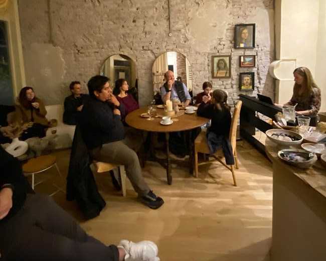 A group of people sits around a table in a cozy setting with exposed brick walls and framed pictures in the background.