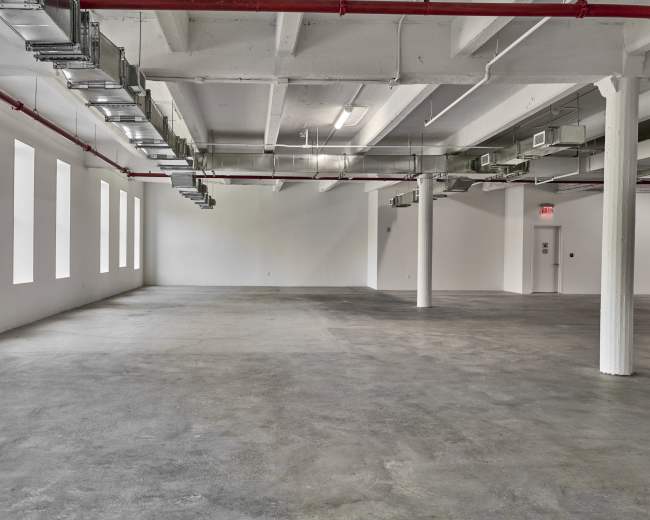 The image shows an empty industrial space with concrete flooring, exposed ductwork, and large windows along the walls.
