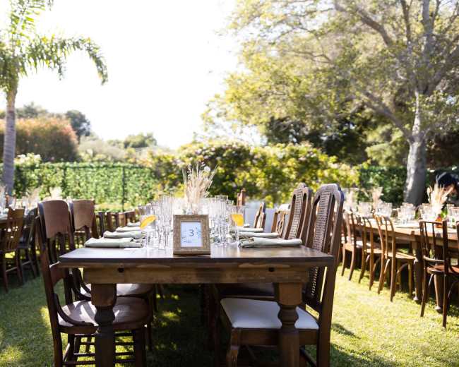 A long outdoor dining setup features wooden tables and chairs, arranged neatly on a grassy area with greenery in the background.