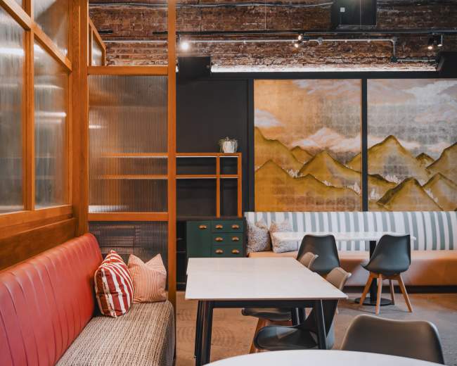 The image shows a modern café interior featuring wooden furniture, a striped sofa, and a mural of mountains on the wall.