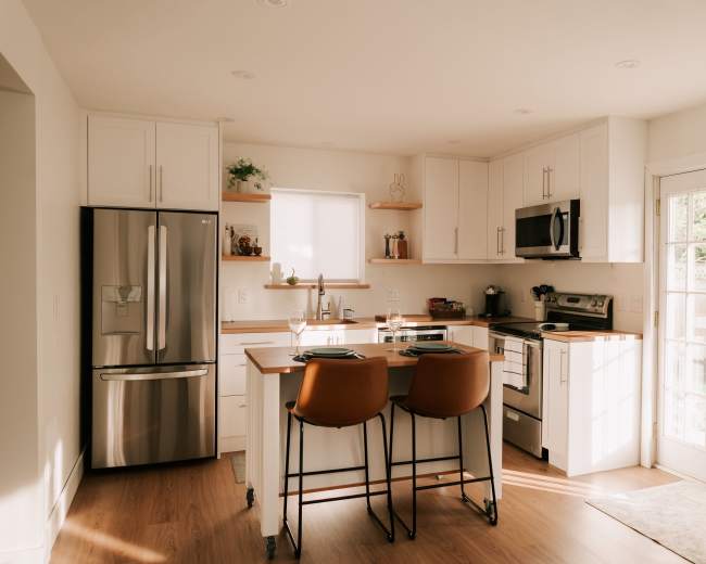 The image shows a modern kitchen featuring white cabinets, stainless steel appliances, a wooden island with two orange bar stools, and a door leading to an outdoor area.