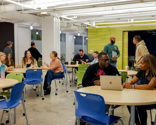 A group of people is engaged in conversations and work at tables in a modern, brightly lit workspace.