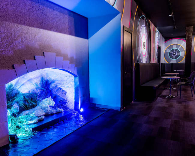 The image features a brightly lit hallway with colorful wall designs, a seating area, and a decorative lion statue surrounded by plants illuminated by blue lighting.
