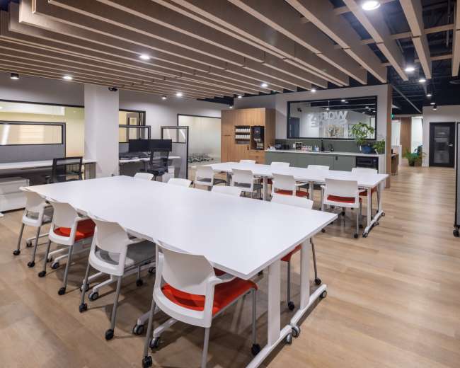The image shows a modern office space featuring multiple white tables with wheels, red-and-white chairs, and a reception area in the background.