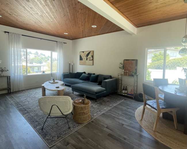 A bright, contemporary living area features a dark sectional sofa, a round coffee table, and a dining area with a wooden table, all set against a backdrop of wooden ceilings and large windows.