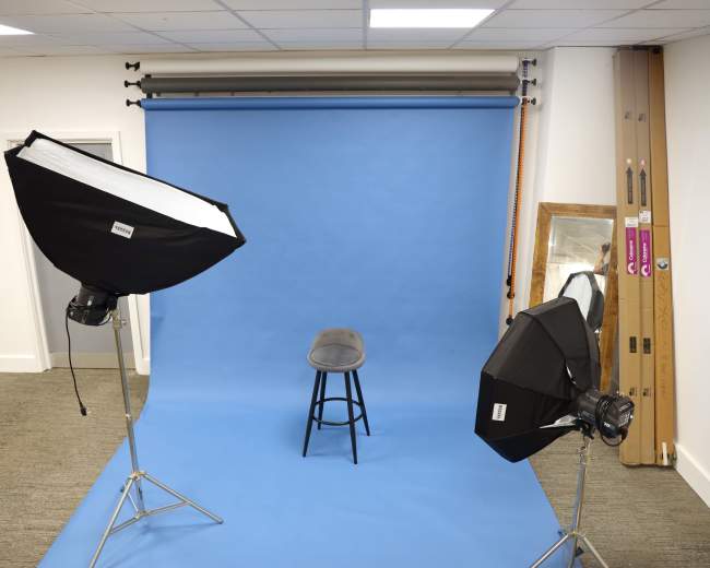 The image shows a photography studio setup with two softbox lights pointing at a single black stool in front of a blue backdrop.