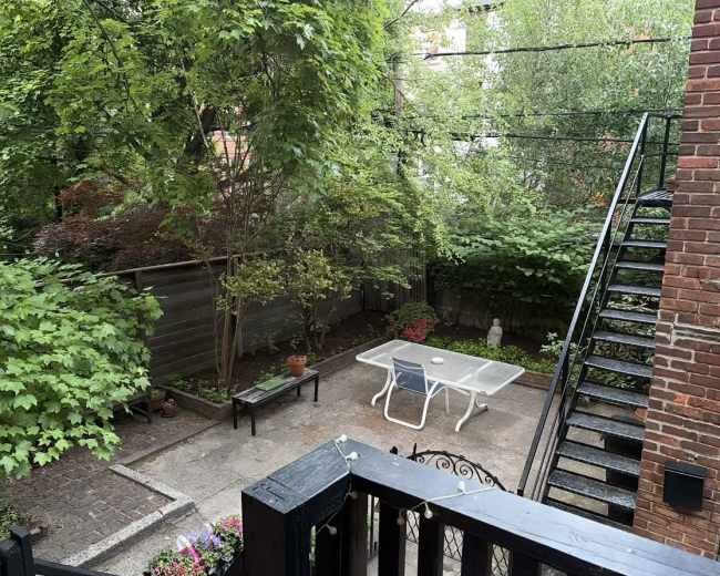 The image shows a small, green backyard with a patio table and chairs surrounded by trees and plants, along with a staircase leading to an upper level.