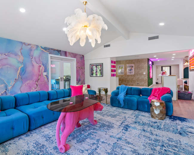 The image showcases a brightly colored living room featuring a blue sectional sofa, a pink table shaped like a lion, and a wall adorned with a pink and purple marbled design.