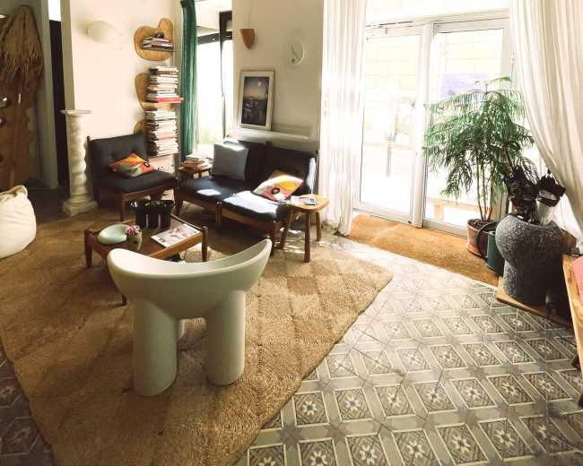 The image shows a cozy living room with a seating area, a large window allowing natural light, and plants adorning the space.