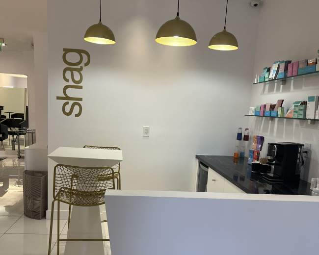 A modern reception area with a white wall, gold pendant lights, and a small counter with a coffee machine and product display shelves.