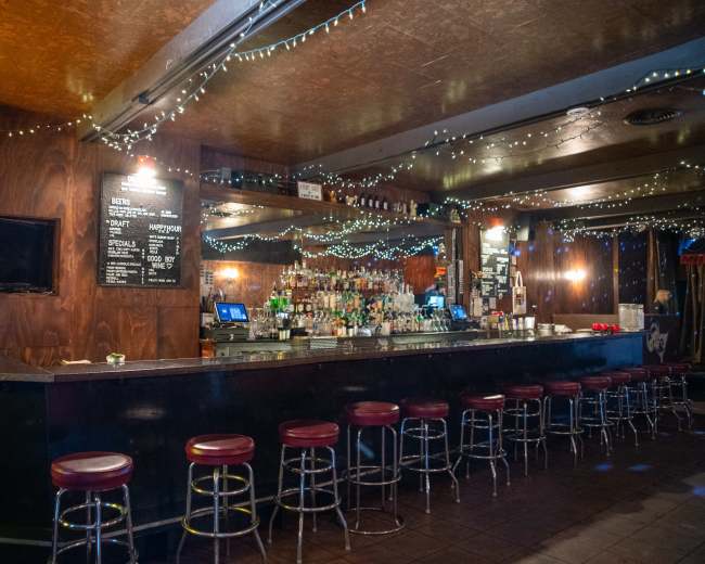 The image shows a dimly lit bar with a long, dark countertop, red stools, and decorative string lights overhead.