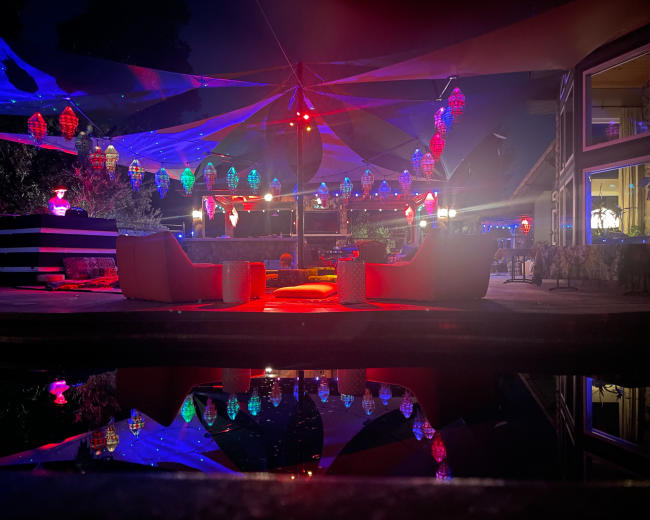 A nighttime patio scene illuminated by colorful lights, featuring two white chairs facing a pool that reflects the vibrant decorations overhead.