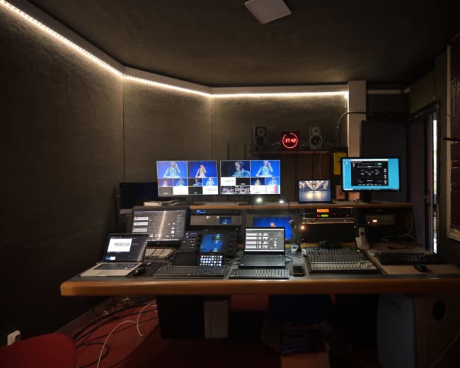The image shows a media control room with multiple screens and equipment arranged on a desk, illuminated by soft ambient lighting.