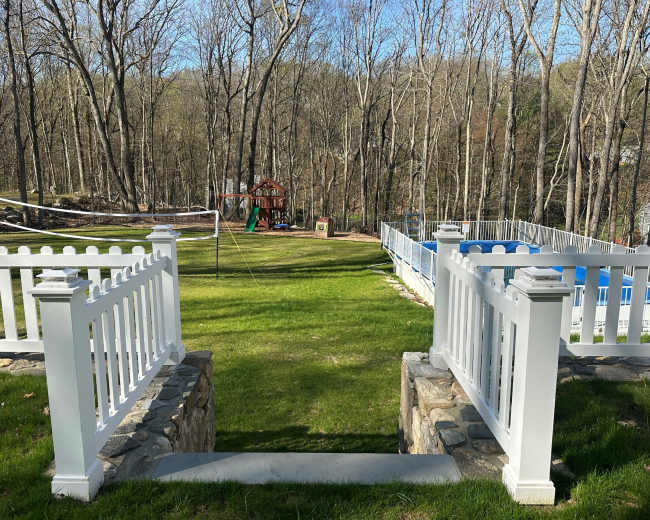 A stone pathway leads down from a white picket fence to a grassy area with a swing set and a play structure, surrounded by trees and a pool.