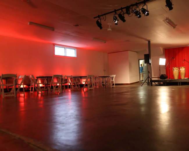 The image shows an indoor event space with red lighting, rows of white folding chairs, and a small stage featuring podiums and musical instruments.