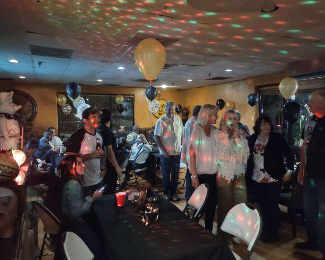 A group of people stands around a decorated room filled with balloons and colorful lights, while others are seated at tables.