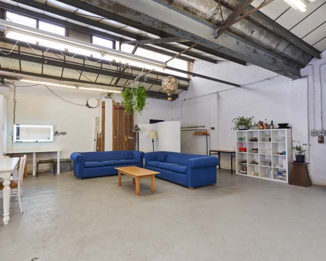 A spacious industrial-style room features two blue sofas, a wooden coffee table, and a dining area with chairs, alongside shelves and a large window.
