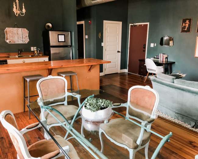 The image shows a modern living space featuring a kitchen bar, dining area with chairs, and a cozy seating area with a couch and decorative elements.