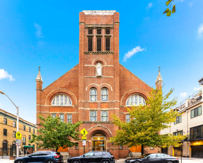 A red brick church with arched windows and a tall bell tower stands against a clear blue sky, flanked by trees and parked cars.