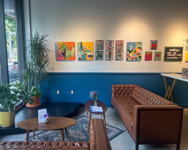 The image shows a contemporary art gallery with colorful paintings displayed on the wall, two brown leather sofas arranged in front of a low table, and plants enhancing the ambiance.