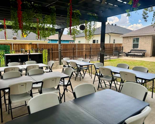 A covered outdoor seating area features multiple rows of folding tables and chairs, with greenery and decorative elements hanging overhead.