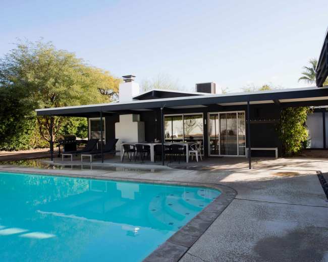 A modern house with a flat roof and large glass windows overlooks a swimming pool surrounded by a concrete patio.