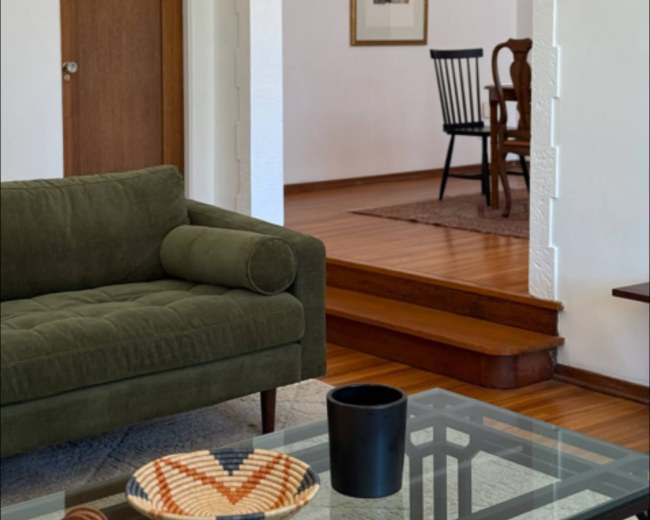 The image shows a living room with a green sofa and an archway leading to a dining area, where a wooden chair and a framed picture are visible.