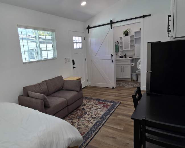 The image shows a small, modern studio space with a bed, a couch, a dining table, and a kitchenette, featuring a sliding barn door and large windows.