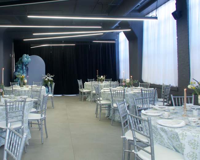 A spacious event room features tables covered with floral tablecloths and set with candles, surrounded by modern silver chairs and decorated with plants and balloons.