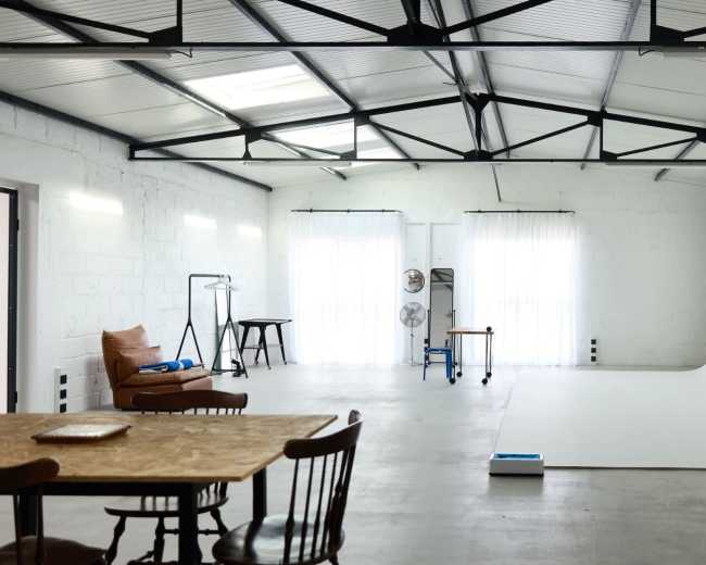 The image shows a spacious photography studio with a white backdrop, natural light streaming through large windows, and a wooden table with chairs.