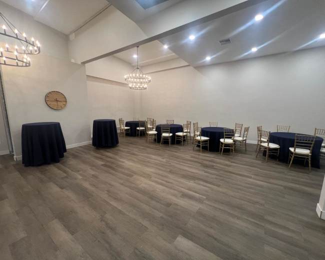 The image shows a spacious event room set with round tables and tall cocktail tables, illuminated by hanging light fixtures.