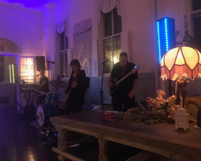 A three-piece band performs on a stage in a dimly lit room with decorative lamps and plants.