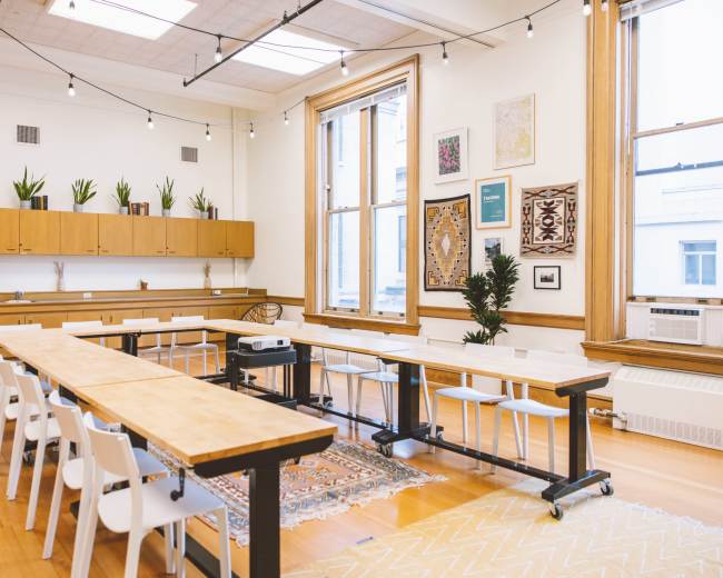 A spacious meeting room features long wooden tables, white chairs, and large windows with plants placed on shelves and along the walls.
