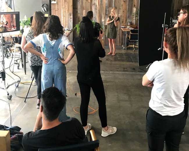 A group of people observes a woman posing for a photoshoot in a studio with wooden walls and various equipment.