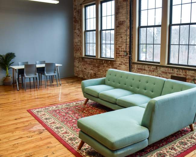The image shows a modern living space with a light blue sectional sofa, a patterned rug, and a dining table with chairs against a brick wall.