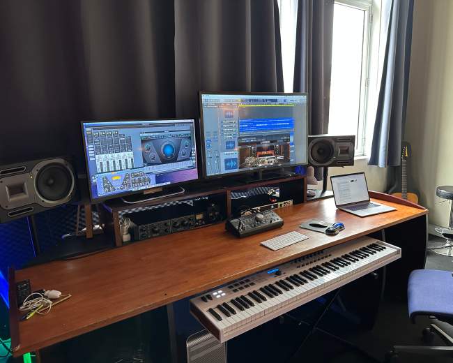 A music production studio is equipped with dual monitors, speakers, a keyboard, and a laptop on a wooden desk.