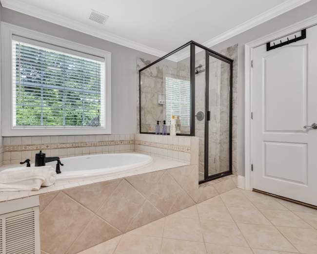 The image shows a bathroom featuring a corner bathtub, a glass-enclosed shower, and a window with blinds.