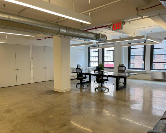 The image shows an empty office space with a large table and four chairs, featuring concrete floors and windows that allow natural light.