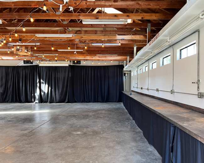 The image shows an indoor event space with wooden beams, string lights, a concrete floor, and a long counter next to a row of windows covered by black curtains.
