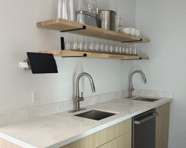 The image shows a modern kitchen countertop with two stainless steel sinks, a dishwasher, and open shelving above displaying glassware and containers.